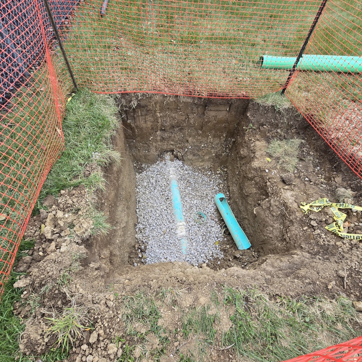 An excavated trench with safety fencing, gravel bedding, and new pipe installed